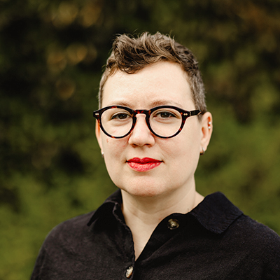 Sasha has short brown hair and is wearing rounded tortoise shell glasses and a black linen button down shirt